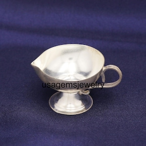 Op de afbeelding: Een kleine, zilverkleurige creamer met een tuit en handvat. De creamer staat op een voetstuk. Het handvat heeft een decoratief ontwerp. De tekst "usagemsjewelry" is zichtbaar op de voorkant van de creamer.