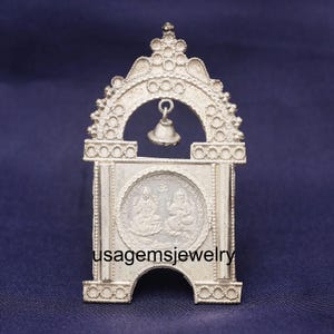 May include: A silver-colored, ornate religious shrine with a bell hanging from the arch. The shrine features a circular design with two figures and the text "usagemsjewelry". The background is a solid dark blue.