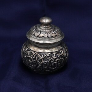 May include: A small, round, silver-toned metal jar with a matching lid. The body features intricate, embossed floral scrollwork patterns, while the lid has a smooth, rounded knob handle and a decorative, scalloped edge design around the top.