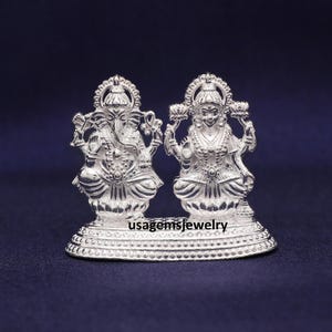 May include: A silver-colored statue featuring two deities, likely Hindu gods, standing side-by-side on a decorative base. The figures are intricately detailed, with ornate clothing and headdresses. The base has the text "usagemsjewelry".