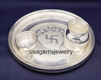 Può includere: Un set thali puja in argento con una piccola ciotola e un piccolo vaso, il tutto su un vassoio rotondo. Il vassoio presenta un disegno in rilievo con un simbolo della svastica. Il testo "usagemsjewelry" è visibile sul vassoio.