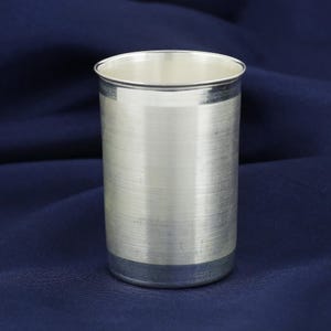 May include: A silver-colored, cylindrical drinking glass with a brushed metal finish. The glass has a simple, elegant design with a slightly flared rim and a band at the base. The background is a dark blue fabric.