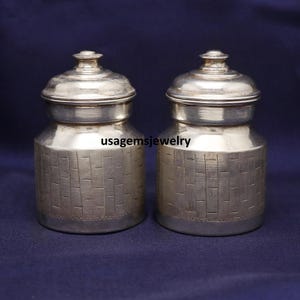 Peut inclure: Deux pots argentés avec couvercles. Les pots sont cylindriques avec un motif texturé en forme de briques au milieu. Les couvercles sont arrondis avec un petit bouton sur le dessus. Le fond est bleu foncé.