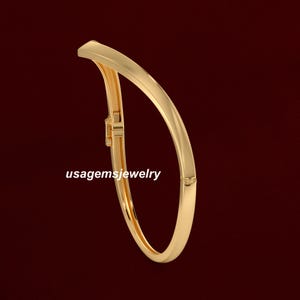 May include: A gold-coloured bangle bracelet with a curved design. The bracelet has a clasp closure and the text "usagemsjewelry" is printed on the bracelet. The jewellery is set against a dark red background.