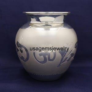 Puede incluir: Una olla redonda de color plateado con boca ancha y diseño decorativo floral y símbolo Om. La olla tiene un acabado pulido y está sobre un fondo azul oscuro. El texto "usagemsjewelry" es visible en la olla.