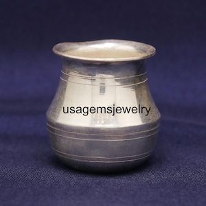 May include: A small, silver-colored metal pot with a rounded body and a flared rim. The pot has horizontal etched lines around its midsection and base. The text "usagemsjewelry" is visible on the front of the pot. The background is a dark blue fabric.