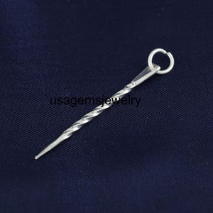 Puede incluir: Una pieza de joyería de metal plateado retorcido con un pequeño lazo en la parte superior y un extremo puntiagudo. El texto "usagemsjewelry" está impreso en el fondo azul oscuro.