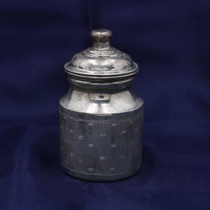 Peut inclure: Un récipient cylindrique en métal argenté avec un couvercle amovible assorti doté d'un bouton de préhension. Le corps du contenant est orné de lignes verticales gravées et de petits tirets horizontaux formant un motif texturé.