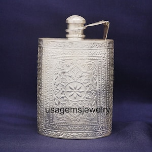 Puede incluir: Petaca plateada con intrincados patrones florales y geométricos en relieve. La petaca tiene una tapa de rosca y un asa con bisagras. El texto "usagemsjewelry" es visible en la parte delantera. La petaca es rectangular y tiene bordes redondeados.