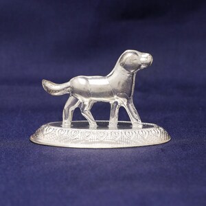 May include: A silver dog ornament on a decorative base. The dog is in a standing position, with its head up. The base features a detailed heart-shaped design. The background is a dark blue. The ornament is likely made of metal.