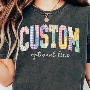 May include: Dark gray t-shirt with the word "CUSTOM" in colorful patchwork letters. Below the word "CUSTOM" is the text "optional line". The shirt is a casual style.