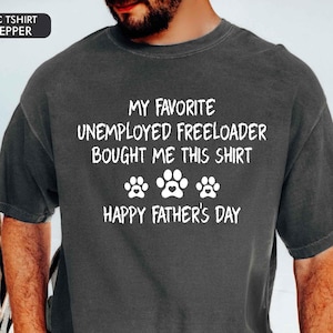 May include: Dark gray t-shirt with white text that reads "MY FAVORITE UNEMPLOYED FREELOADER BOUGHT ME THIS SHIRT" above three paw prints and "HAPPY FATHER'S DAY". The shirt is a casual style.
