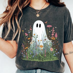 May include: Dark gray t-shirt featuring a white ghost with black eyes and a floral meadow design. The ghost is surrounded by colorful flowers and butterflies. The shirt has short sleeves and a crew neck.