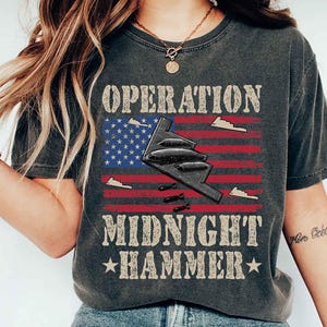 May include: Dark gray t-shirt with the text "OPERATION MIDNIGHT HAMMER" above and below a graphic of an American flag and a stealth bomber. The flag has red stripes and a blue field with white stars.