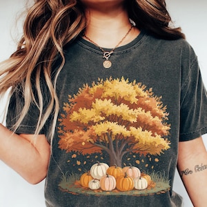 May include: Dark gray t-shirt featuring a fall-themed graphic. The design includes a tree with yellow and orange leaves, pumpkins, and fallen leaves. The shirt has a vintage, worn look.