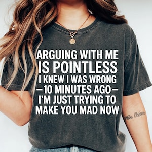 May include: Dark gray t-shirt with the text "ARGUING WITH ME IS POINTLESS I KNEW I WAS WRONG -10 MINUTES AGO- I'M JUST TRYING TO MAKE YOU MAD NOW" in white. The shirt is a casual wear item.