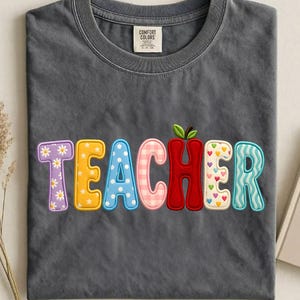 May include: A gray Comfort Colors t-shirt with the word "TEACHER" in colorful, embroidered letters. The letters feature various patterns, including flowers, polka dots, and hearts. An apple graphic replaces the letter "A".