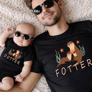 May include: Matching black t-shirts and a onesie featuring otter illustrations. The adult's shirt says "FOTTER" and the baby's onesie says "DOTTER". Both shirts have a brown otter family design with a heart.