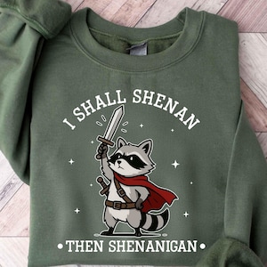May include: Green sweatshirt with the text "I SHALL SHENAN... THEN SHENANIGAN." Features a cartoon raccoon holding a sword and wearing a red cape. The raccoon has a black mask and a striped tail.