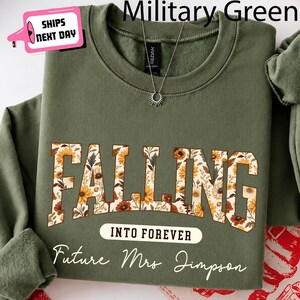 May include: A military green crewneck sweatshirt featuring the word FALLING in large block letters filled with a floral pattern of sunflowers and autumn leaves, followed by the text INTO FOREVER and Future Mrs Jimpson in white script.