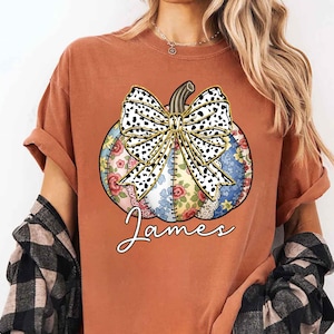 May include: Burnt orange t-shirt featuring a colorful pumpkin design with a dalmatian print bow and floral patchwork. The name "James" is written in cursive below the pumpkin. The shirt has rolled-up sleeves and a relaxed fit.