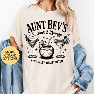 May include: Ivory-colored t-shirt with a black graphic that reads "Aunt Bev's Sidebar & Lounge" and features illustrations of cocktails. The text "Stay Salty, Resist Often" is below the illustrations. The shirt has a round neck and short sleeves.