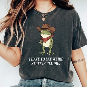 May include: Dark gray t-shirt featuring a cartoon frog wearing a cowboy hat and red bandana, holding a coffee cup. The text "I HAVE TO SAY WEIRD STUFF IR I'LL DIE" is printed below the frog in white.