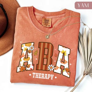 May include: A terracotta-colored t-shirt with the text "ABA THERAPY" in a collegiate-style font. The letters are decorated with fall leaves, pumpkins, and daisies. A gold necklace with a pendant is draped across the shirt.