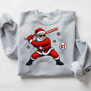 May include: A light gray sweatshirt featuring a graphic of Santa Claus in a baseball stance, swinging a candy cane bat. Santa is wearing sunglasses and a red suit. A baseball with the number 67 is in the image.
