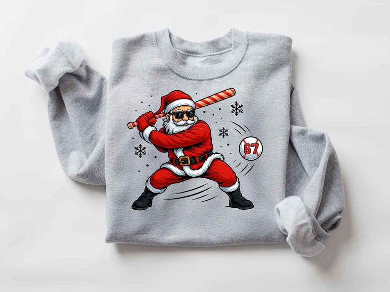 May include: A light gray sweatshirt featuring a graphic of Santa Claus in a baseball stance, swinging a candy cane bat. Santa is wearing sunglasses and a red suit. A baseball with the number 67 is in the image.