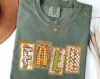 May include: Sage green t-shirt with the word "FALL" in a decorative design. The letters are in individual frames with fall-themed patterns, including pumpkins, plaid, and floral prints. A silver necklace with a pendant is also visible.