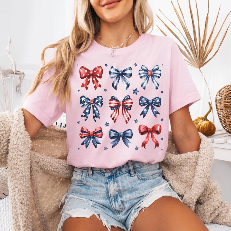 May include: A pink t-shirt with a pattern of red, white, and blue bows. The bows are arranged in a grid pattern and are all facing the same direction. The shirt is made of a soft, comfortable material and is perfect for everyday wear.