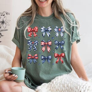 May include: A green t-shirt with a pattern of red, white, and blue bows. The bows are arranged in a grid pattern and are all different sizes. The shirt is made of a soft, comfortable material and is perfect for everyday wear.