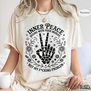 Puede incluir: Camiseta color crema con el texto "INNER PEACE BEGINS WITH FOUR WORDS: NOT MY F*CKING PROBLEM" en fuente g&oacute;tica negra. El dise&ntilde;o incluye una mano de esqueleto haciendo el signo de la paz, rodeada de elementos florales.