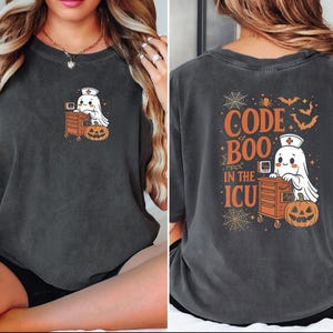 May include: Dark gray t-shirt with a Halloween-themed design. The front features a cartoon ghost wearing a nurse's cap, a medical cart, and a jack-o'-lantern. The back of the shirt has the text "CODE BOO IN THE ICU" with bats and spiderwebs.
