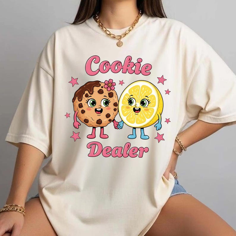 Puede incluir: Camiseta color crema con una galleta y un lim&oacute;n de dibujos animados tomados de la mano, con el texto "Cookie Dealer" en rosa. La galleta tiene chispas de chocolate y una flor, mientras que el lim&oacute;n tiene una cara sonriente. Estrellas rosas rodean los personajes.