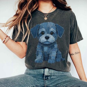 May include: Dark gray t-shirt featuring a blue denim patchwork puppy design. The graphic shows a seated dog with large eyes and a denim collar. The shirt is a casual style, suitable for everyday wear.