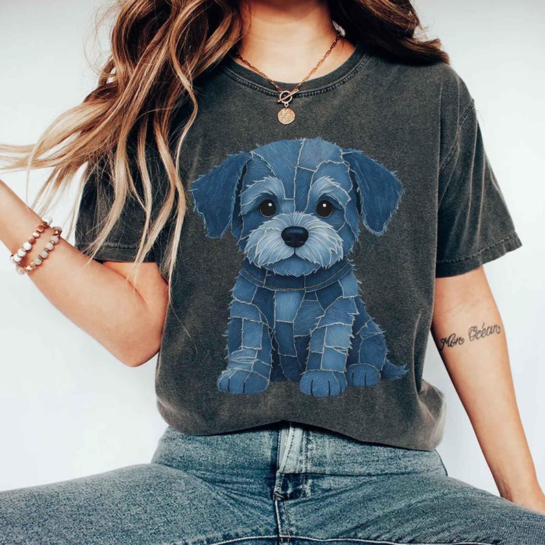 May include: Dark gray t-shirt featuring a blue denim patchwork puppy design. The graphic shows a seated dog with large eyes and a denim collar. The shirt is a casual style, suitable for everyday wear.