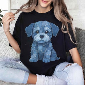 May include: Black t-shirt featuring a blue denim-style puppy illustration. The puppy is sitting, with a light blue nose and white accents. The design is centered on the shirt.