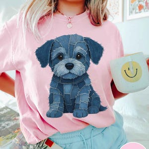 May include: Pink crew neck sweatshirt featuring a denim-style puppy graphic. The puppy is blue and has a patchwork design. A white mug with a yellow smiley face is held in the background.