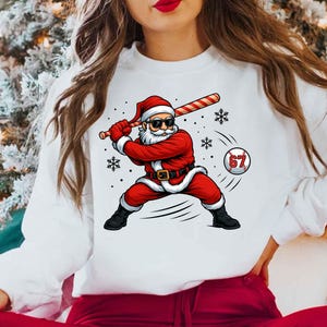 May include: White sweatshirt featuring a graphic of Santa Claus in a baseball stance, swinging a bat. Santa is wearing a red suit, hat, and sunglasses. A baseball with the number 67 is in the image, along with snowflakes.