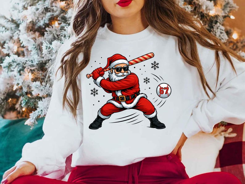 May include: White sweatshirt featuring a graphic of Santa Claus in a baseball stance, swinging a bat. Santa is wearing a red suit, hat, and sunglasses. A baseball with the number 67 is in the image, along with snowflakes.