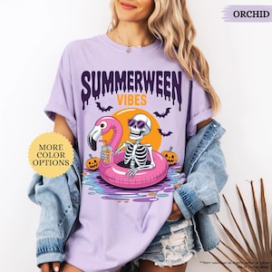 K&ouml;nnte beinhalten: Orchideenfarbenes lila T-Shirt mit einem Grafikdesign, das ein Skelett mit Sonnenbrille auf einem rosa Flamingo-Schwimmring zeigt. Das Shirt hat die Aufschrift "SUMMERWEEN VIBES", Flederm&auml;use, K&uuml;rbisse und eine Sonne.