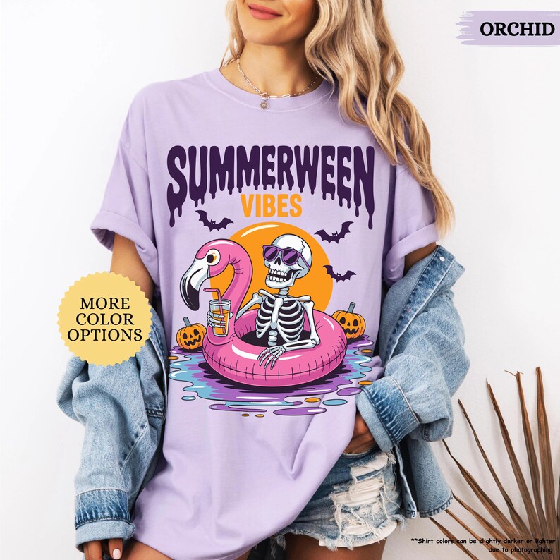 K&ouml;nnte beinhalten: Orchideenfarbenes lila T-Shirt mit einem Grafikdesign, das ein Skelett mit Sonnenbrille auf einem rosa Flamingo-Schwimmring zeigt. Das Shirt hat die Aufschrift "SUMMERWEEN VIBES", Flederm&auml;use, K&uuml;rbisse und eine Sonne.