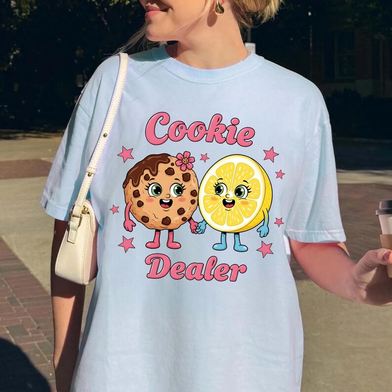 Puede incluir: Camiseta azul claro con un dise&ntilde;o l&uacute;dico que presenta una galleta y un lim&oacute;n sonrientes tomados de la mano, con el texto "Cookie Dealer" en rosa. El dise&ntilde;o incluye estrellas rosas y una flor en la galleta.