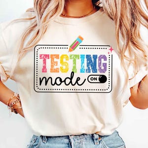 May include: A white t-shirt with a rainbow colored graphic that says "TESTING mode ON" with a pencil icon above the text.