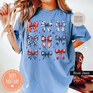 May include: A blue t-shirt with a red, white, and blue bow design. The shirt has the text "Comfort Colors" on a white tag and "Ships Next Day" on a pink tag.