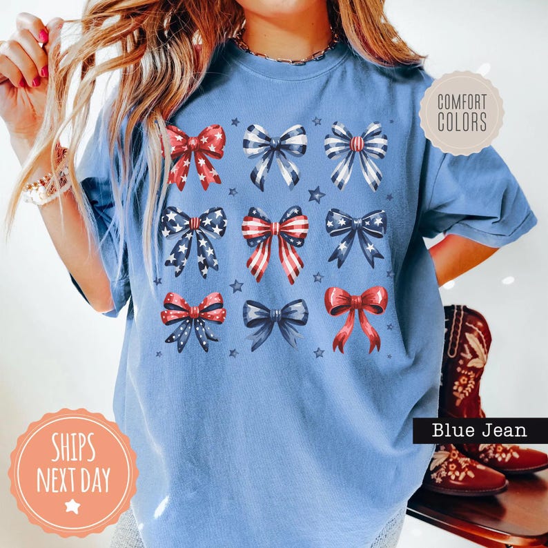 May include: A blue t-shirt with a red, white, and blue bow design. The shirt has the text "Comfort Colors" on a white tag and "Ships Next Day" on a pink tag.