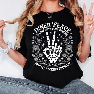 Puede incluir: Camiseta negra con un gr&aacute;fico blanco que presenta una mano de esqueleto haciendo el signo de la paz. El texto dice "INNER PEACE BEGINS WITH FOUR WORDS: NOT MY F*CKING PROBLEM" rodeado de dise&ntilde;os florales y estrellas.