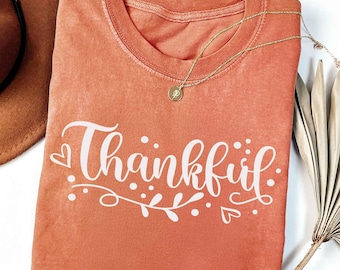 May include: An orange-colored t-shirt with the word "Thankful" in white script, adorned with decorative leaves and hearts. A gold necklace with a pendant rests on the shirt. A brown hat and dried palm leaves are partially visible.
